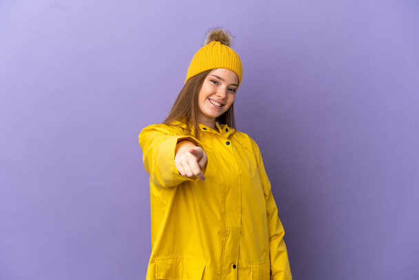 Ragazza adolescente che indossa un cappotto antipioggia su sfondo viola isolato che punta davanti con espressione felice - Foto, immagini