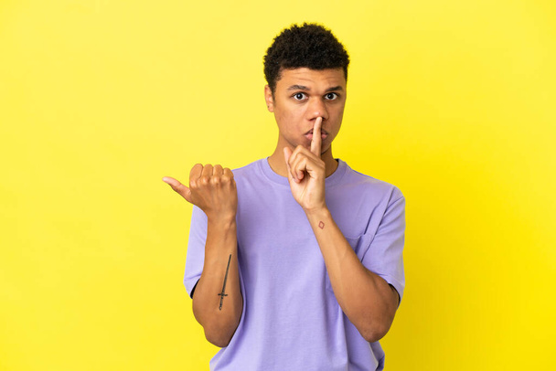 Young African American man isolated on yellow background pointing to the side and doing silence gesture - 写真・画像