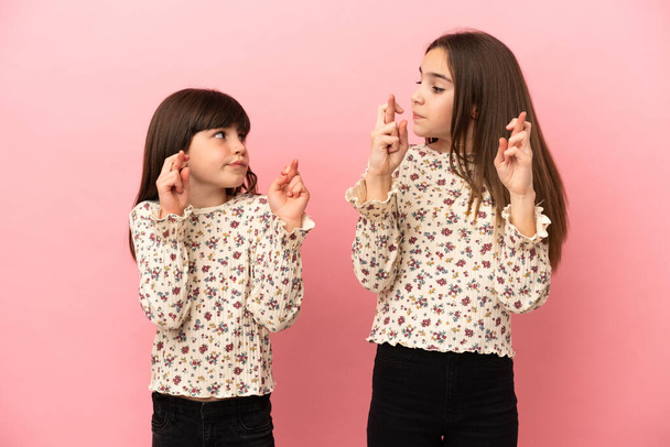 Piccole sorelle ragazze isolate su sfondo rosa con le dita incrociate e augurando il meglio - Foto, immagini