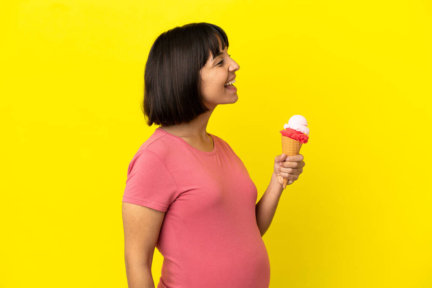 Pregnant woman holding a cornet ice cream isolated on yellow background laughing in lateral position - Photo, Image
