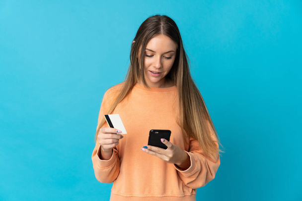 Jeune femme lituanienne isolée sur fond bleu achetant avec le mobile avec une carte de crédit - Photo, image