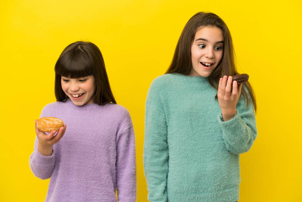 Little sisters isolated on yellow background holding donuts with happy expression - Foto, immagini