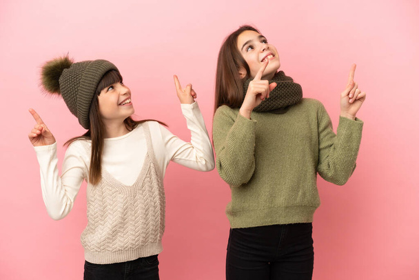 Malé sestry v zimním oblečení izolované na růžovém pozadí ukazuje ukazováčkem skvělý nápad - Fotografie, Obrázek