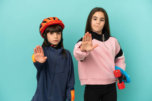 Little sisters practicing cycling and skater isolated on blue background making stop gesture denying a situation that thinks wrong - Photo, Image