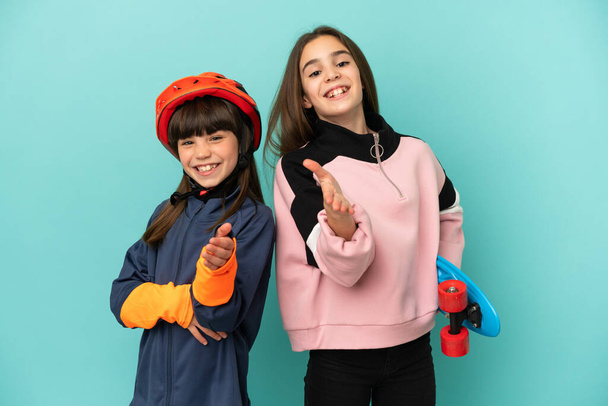 Kleine Schwestern üben Radfahren und Skater isoliert auf blauem Hintergrund schütteln Hände, um ein gutes Geschäft zu schließen - Foto, Bild