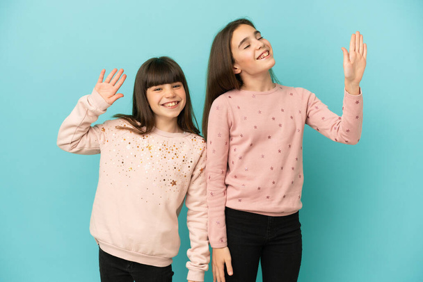 Siostry dziewczęta odizolowane na niebieskim tle salutując ręką ze szczęśliwą ekspresją - Zdjęcie, obraz