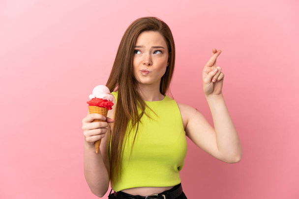 Chica adolescente con un helado de corneta sobre fondo rosa aislado con los dedos cruzando y deseando lo mejor - Foto, Imagen