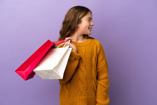 Little caucasian girl isolated on purple background holding shopping bags and looking back - Foto, Imagem