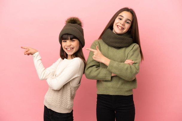 Hermanitas vestidas con ropa de invierno aisladas sobre fondo rosa apuntando con el dedo hacia un lado en posición lateral - Foto, Imagen