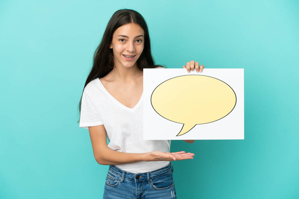 Young French woman isolated on blue background holding a placard with speech bubble icon and pointing it - Foto, imagen