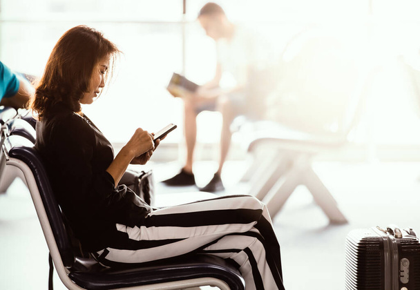 Vrouw zit in de lobby van de luchthaven met behulp van een smartphone en op het scherm te kijken tijdens het wachten op doorvoer. Zonlicht schijnt van buitenaf. - Foto, afbeelding