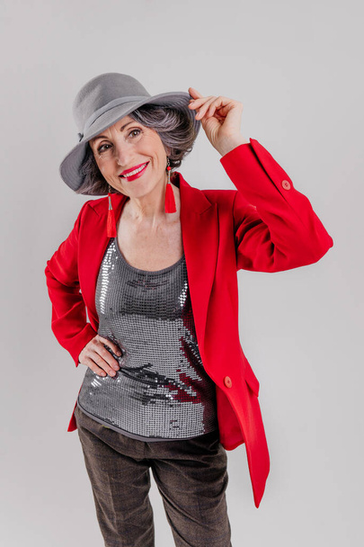 Stylish senior woman wearing red blazer and grey hat on grey background - Zdjęcie, obraz