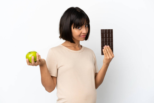 Молода змішана раса вагітна жінка ізольована на білому тлі, приймаючи шоколадну таблетку в одній руці і яблуко в іншій
 - Фото, зображення