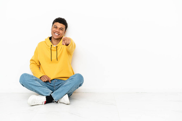 Junger afroamerikanischer Mann, der isoliert auf weißem Hintergrund auf dem Boden sitzt, zeigt mit selbstbewusstem Gesichtsausdruck auf dich - Foto, Bild