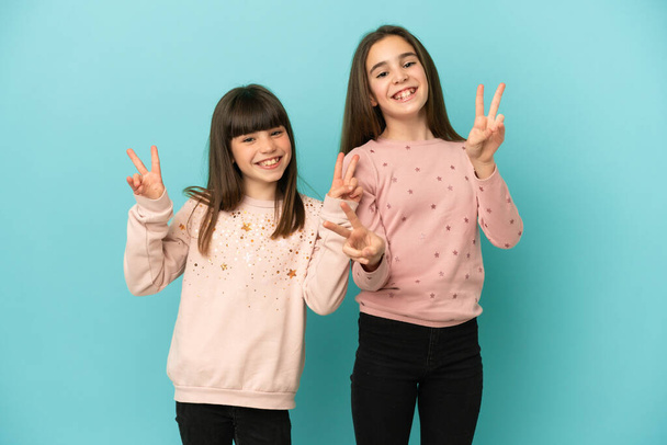 kleine zusjes meisjes geïsoleerd op blauwe achtergrond glimlachen en tonen overwinning teken met beide handen - Foto, afbeelding