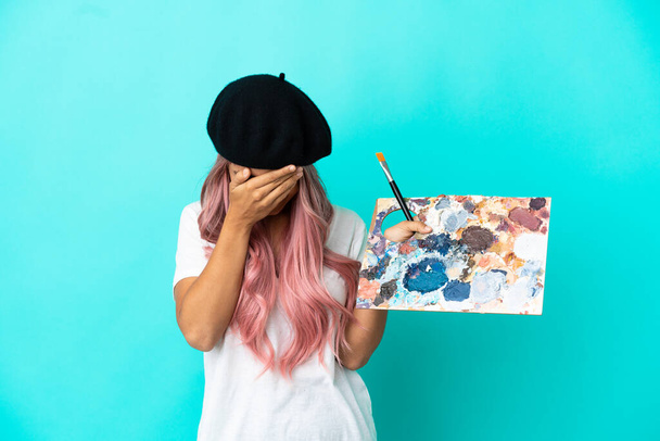 Mladá smíšená rasa žena s růžovými vlasy drží paletu izolované na modrém pozadí s unaveným a nemocným výrazem - Fotografie, Obrázek