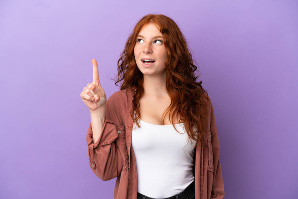 Tini vörös hajú lány felett elszigetelt lila háttér gondolkodás egy ötlet rámutatva az ujját fel - Fotó, kép