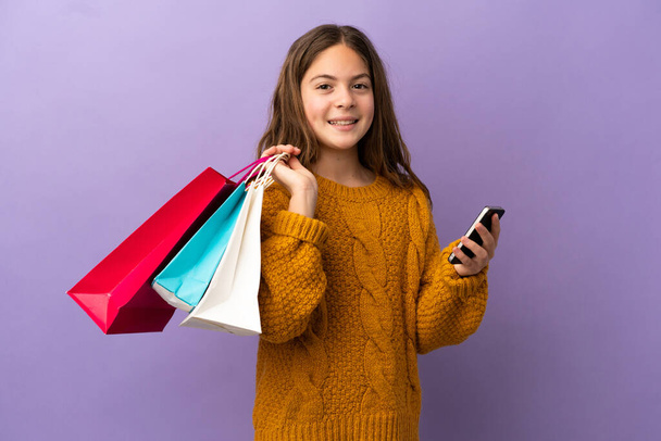 Little caucasian girl isolated on purple background holding shopping bags and writing a message with her cell phone to a friend - Foto, Imagem