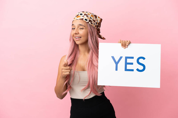 Young mixed race woman with pink hair isolated on pink background holding a placard with text YES and pointing to the front - Photo, Image