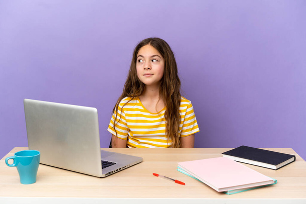 Kleines studentisches Mädchen an einem Arbeitsplatz mit einem Laptop isoliert auf lila Hintergrund macht Zweifel Geste Blick Seite - Foto, Bild