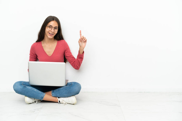 Jovencita francesa sentada en el suelo con su portátil mostrando y levantando un dedo en señal de la mejor - Foto, Imagen