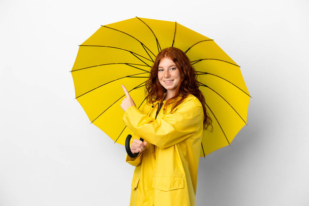Teenager redhead girl rainproof coat and umbrella isolated on white background pointing back - Fotó, kép