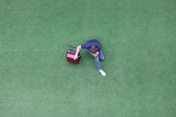 Triste hombre con la maleta sentada en la hierba verde y sosteniendo su cabeza vista superior - Foto, imagen