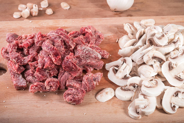 In reepjes gesneden stukken rundvlees, uitgebeend in meel en gehakte champignons op een snijplank, close-up - ingrediënten voor stroganoff van rundvlees met champignons - Foto, afbeelding