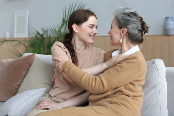 Beautiful mother and daughter. Cheerful young woman is embracing her middle aged mother in living room. Family portrait - Foto, Bild
