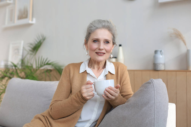 Happy mature woman resting on comfortable sofa drink coffee or tea, looking away, relaxing on cozy couch at home enjoy hot beverage - Фото, изображение