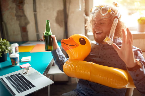 A young man enjoys imagining a beautiful beach while sitting in a relaxed atmosphere in the office with beer and beach accessories - Фото, изображение