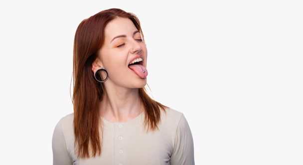 Эмоции женщина показывает язык. С серьгами в носу, торчащими из языка и пирсингом. Белый фон, пространство для копирования. Пирсинг лица женщины - Фото, изображение