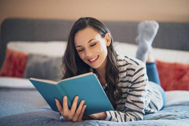 Onnellinen nuori nainen makaa sängyssä kotonaan ja lukee kirjaa. - Valokuva, kuva