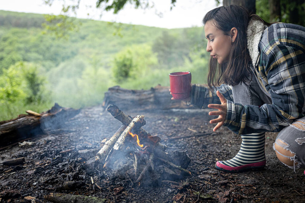 An attractive girl with a cup in her hand warms up near a fire in the forest. - Foto, Bild