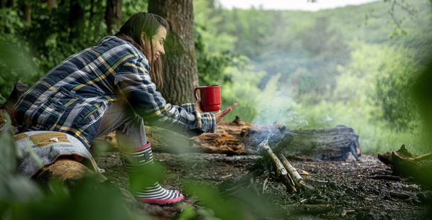 Een aantrekkelijk meisje met een kopje in haar hand warmt op bij een vuur in het bos. - Foto, afbeelding