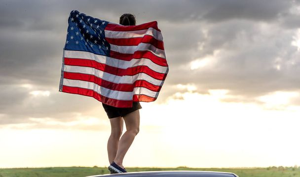 自然の中で夕日を楽しむアメリカ合衆国の国旗を持つ若い女性. - 写真・画像