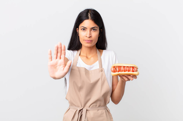hispanic pretty woman pretty hispanic chef woman looking serious showing open palm making stop gesture and holding a hot dog - Foto, Bild