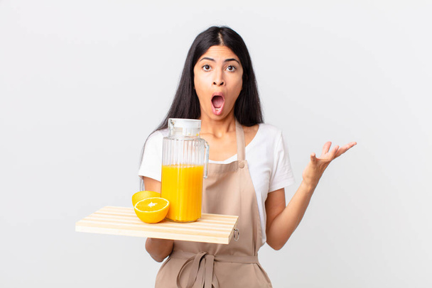 pretty hispanic chef woman feeling extremely shocked and surprised and holding a orange juice - Foto, imagen