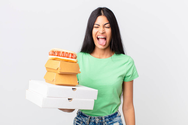pretty hispanic woman with cheerful and rebellious attitude, joking and sticking tongue out and holding take away fast food boxes - Foto, imagen