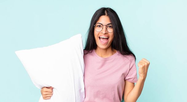 pretty hispanic woman feeling shocked,laughing and celebrating success and wearing pajamas with a pillow - Fotó, kép