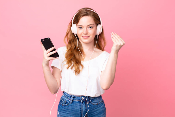 pretty red head woman making capice or money gesture, telling you to pay with headphones and smartphone - Fotografie, Obrázek