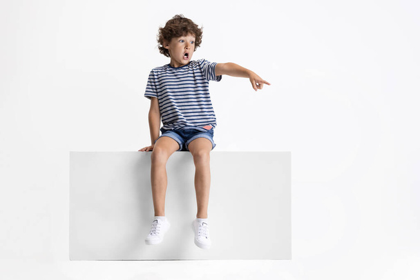 Little Caucasian preschool boy sitting on box isolated over white studio background. Copyspace. Childhood, education, emotions concept - Photo, image