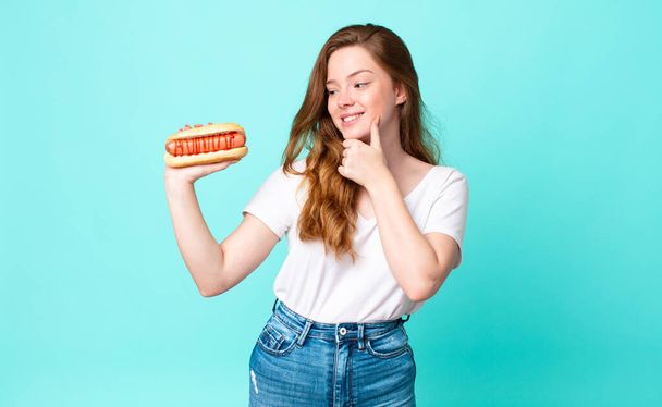 red head pretty woman smiling with a happy, confident expression with hand on chin and holding a hot dog - Foto, Imagem