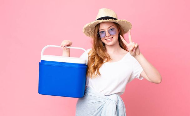 pretty red head woman smiling and looking happy, gesturing victory or peace and holding a picnic portable refrigerator - Fotoğraf, Görsel