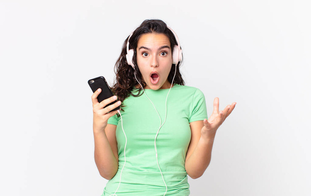 pretty hispanic woman amazed, shocked and astonished with an unbelievable surprise with headphones and a smartphone - Foto, Bild