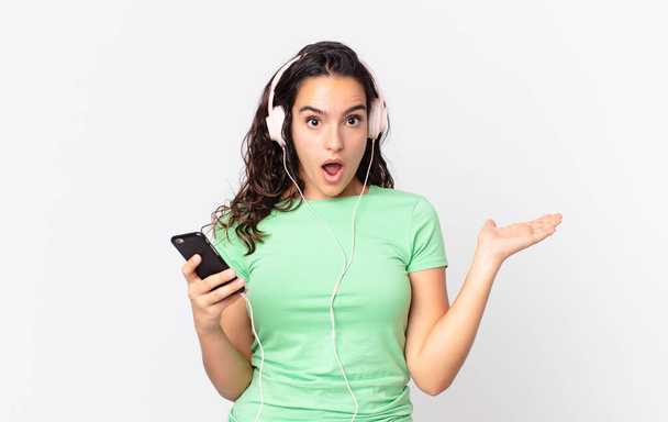 pretty hispanic woman looking surprised and shocked, with jaw dropped holding an object with headphones and a smartphone - Foto, afbeelding