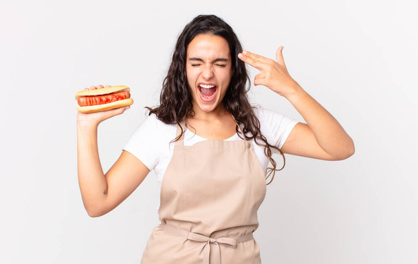 hispanic pretty chef woman looking unhappy and stressed, suicide gesture making gun sign and holding a hot dog - Foto, Bild