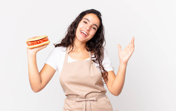 hispanic pretty chef woman feeling happy, surprised realizing a solution or idea and holding a hot dog - Fotó, kép