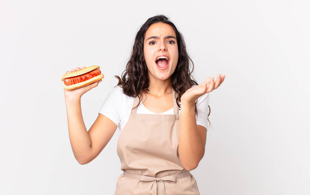 hispanic pretty chef woman looking desperate, frustrated and stressed and holding a hot dog - Foto, Imagem