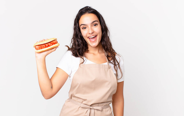 hispanic pretty chef woman looking happy and pleasantly surprised and holding a hot dog - Foto, Bild